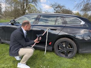 Kfz-Sachverständiger Andreas Anzer fotografiert mit einer Spiegelreflexkamera einen Unfallschaden an der Fahrzeugtür eines schwarzen BMW Kombis – mit Maßstab zur präzisen Schadendokumentation.
