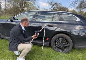 Kfz-Sachverständiger Andreas Anzer fotografiert mit einer Spiegelreflexkamera einen Unfallschaden an der Fahrzeugtür eines schwarzen BMW Kombis – mit Maßstab zur präzisen Schadendokumentation.